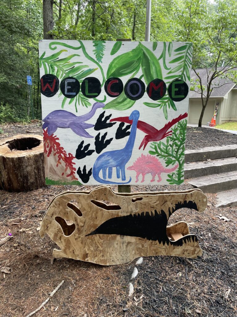 welcome sign with images of dinosaurs, dinosaur tracks, erupting volcano and leaves, with a wooden dinosaur skull in front of it